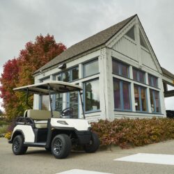 Front-side view of the Toro Vista 4-seater golf cart with beige seats, parked outside a rustic building with fall foliage and cloudy skies