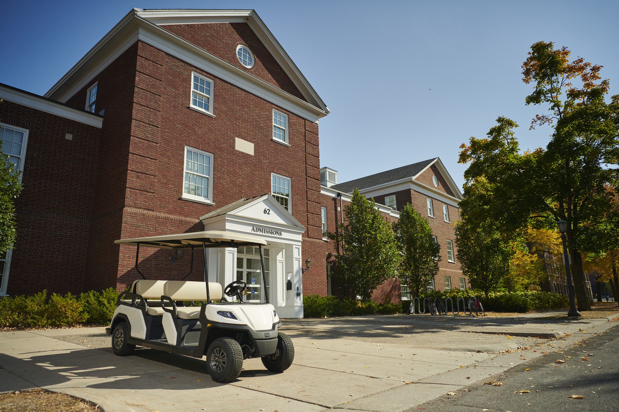 Toro Vista electric golf cart in front of a traditional red-brick college admissions building, surrounded by trees and bike racks.