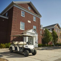 Toro Vista 6-seater golf cart with beige seats, parked outside a university admissions building, surrounded by brick architecture and greenery