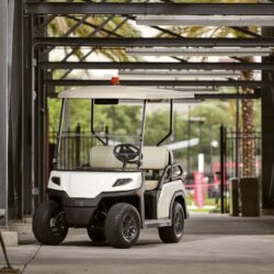 Toro Vista 4-seater golf cart parked in a shaded area with beige seating and a white roof, captured under a structure with metal beams and greenery in the background