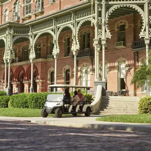 Toro Vista 8-seater golf cart with beige seats and a white roof, transporting passengers past a historic brick building with intricate architectural details