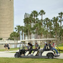 Toro Vista 8-seater golf cart with passengers driving along a sidewalk in a park, with palm trees, skyscrapers, and a river in the background