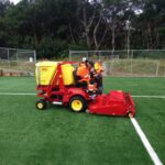 Maintenance crew using SMG SportChamp SC2D ride-on machine to clean artificial turf on a soccer field.