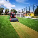 SMG TurfKing TK1502 turf cleaner attached to a red tractor cleaning an artificial soccer field under a clear sky.