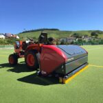 SMG TurfKing TK1502 turf cleaner attached to a tractor on an artificial soccer field with rolling green hills in the background.