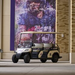 Toro Vista 4-seater golf cart with TCU decals, beige seats, and a white roof, parked near a football-themed mural outside a brick building